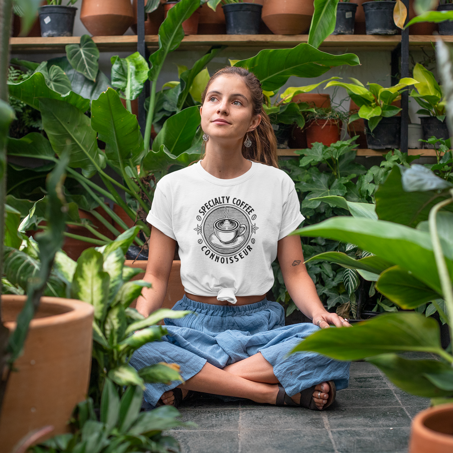 Woman sitting among plants wearing a white t-shirt with a coffee graphic.