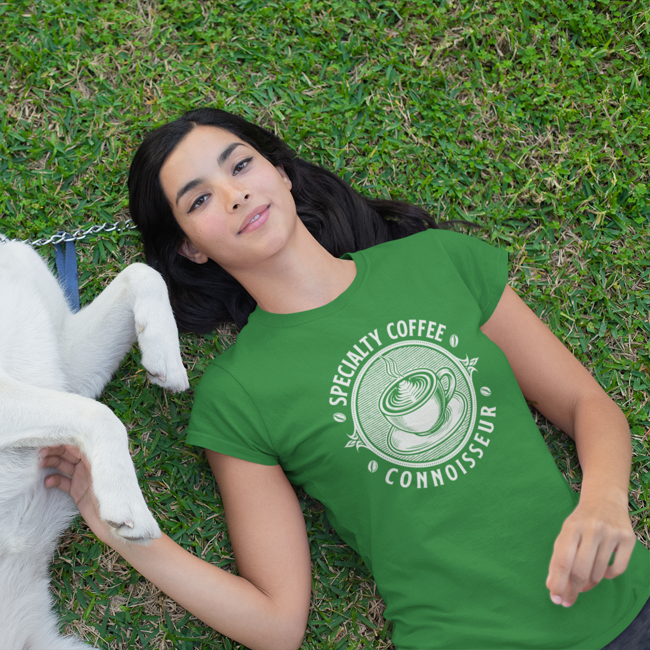 Woman wearing a green t-shirt with a coffee-themed design lying on grass with a white dog.